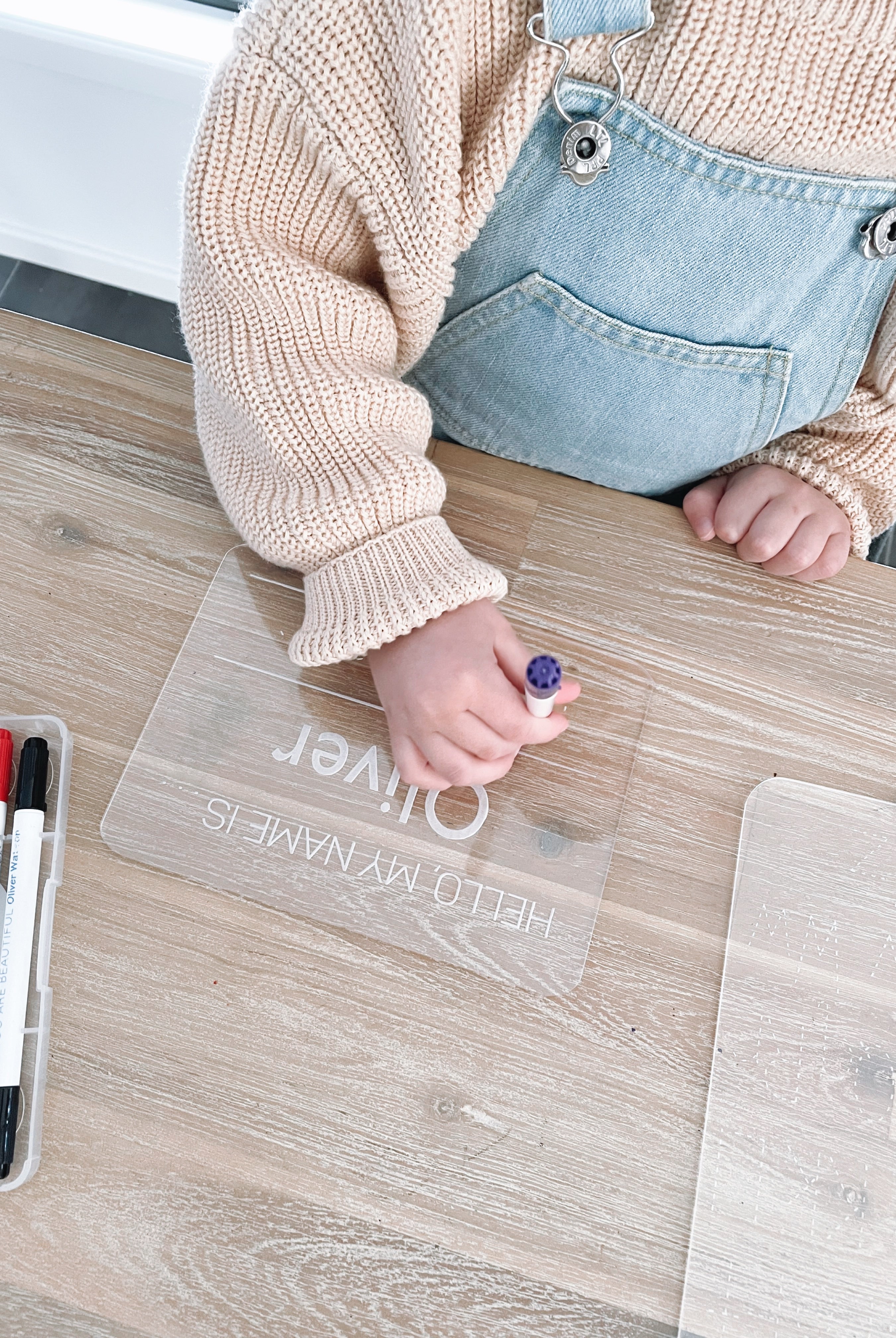 A child interacting with a clear acrylic tracing board with dotted lines, placed on a wooden floor, with markers and a wet wipe in the surrounding area.