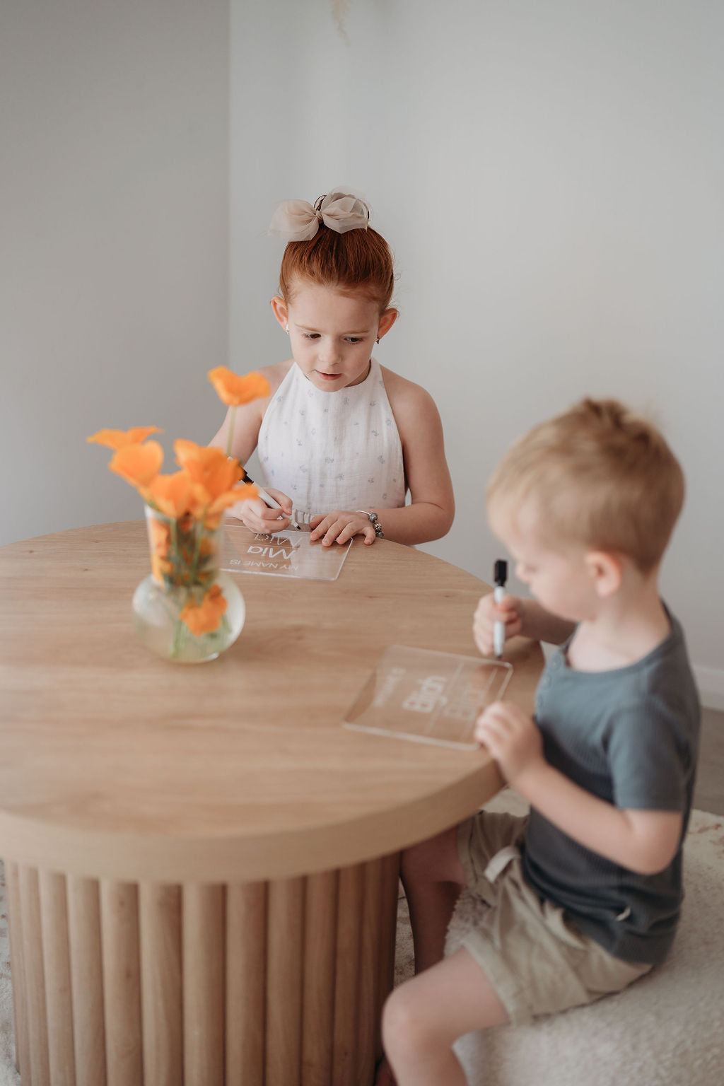 Two children sitting at a wooden table with a vase of flowers and books with name tracing boards