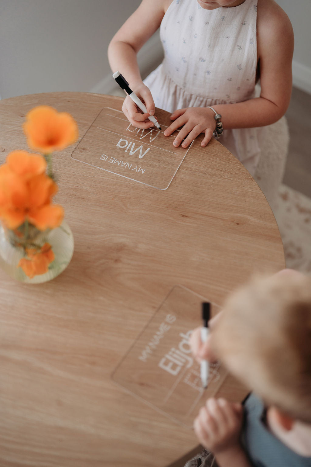 Person writing on clear acrylic name cards with a child nearby, on a wooden table with orange flowers.