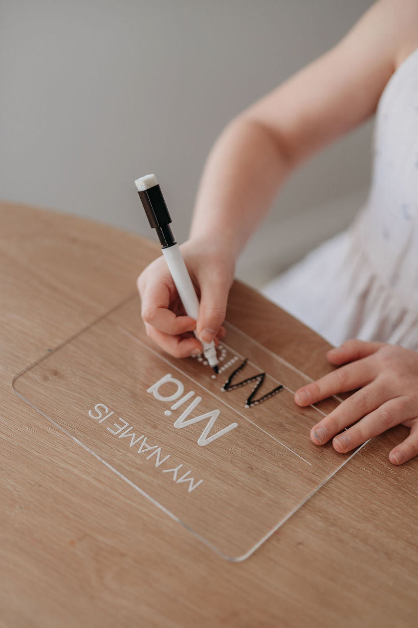 Child writing on a clear acrylic sheet with a pen on a wooden table.