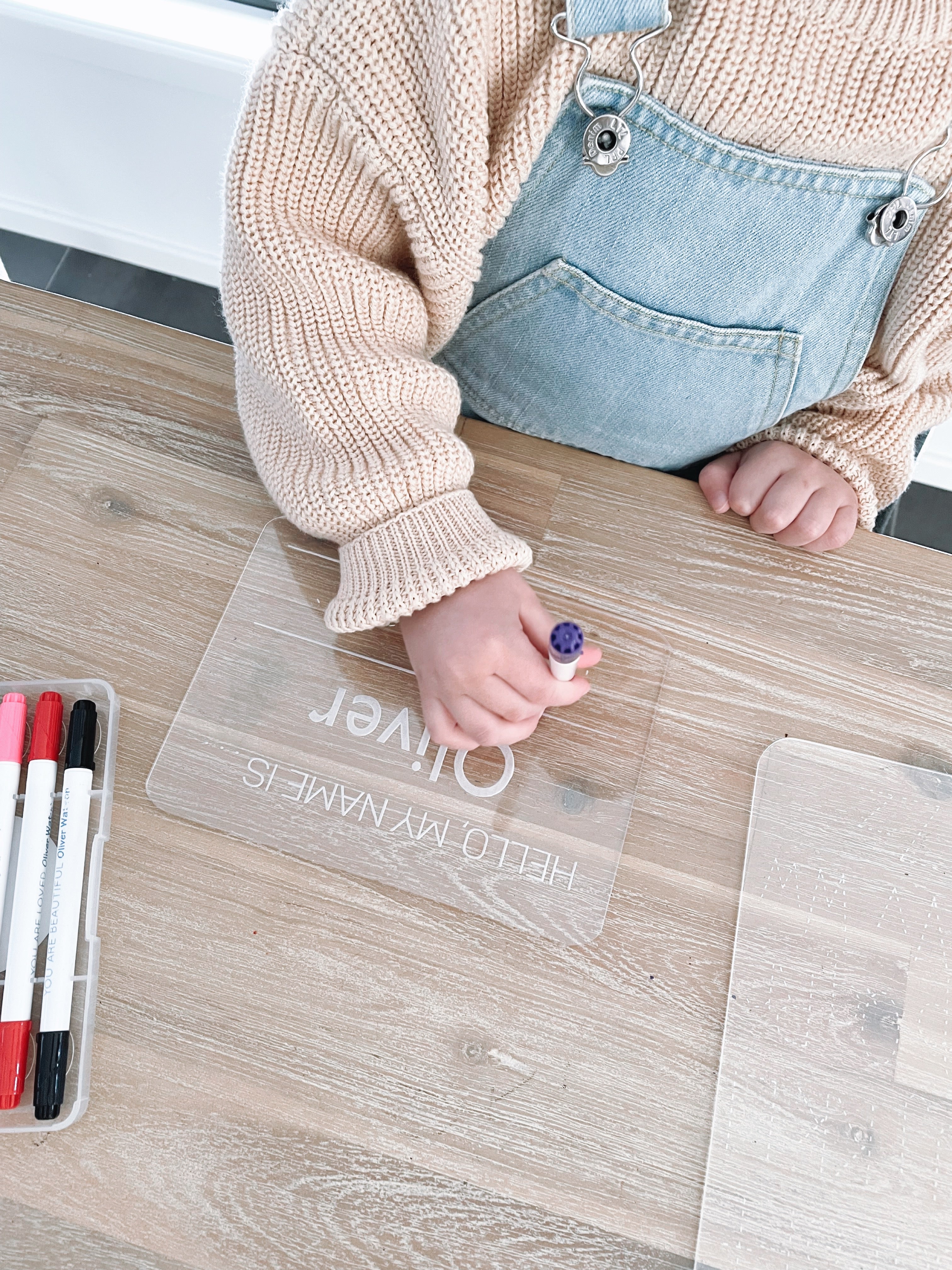 A child interacting with a clear acrylic tracing board with dotted lines, placed on a wooden floor, with markers and a wet wipe in the surrounding area.